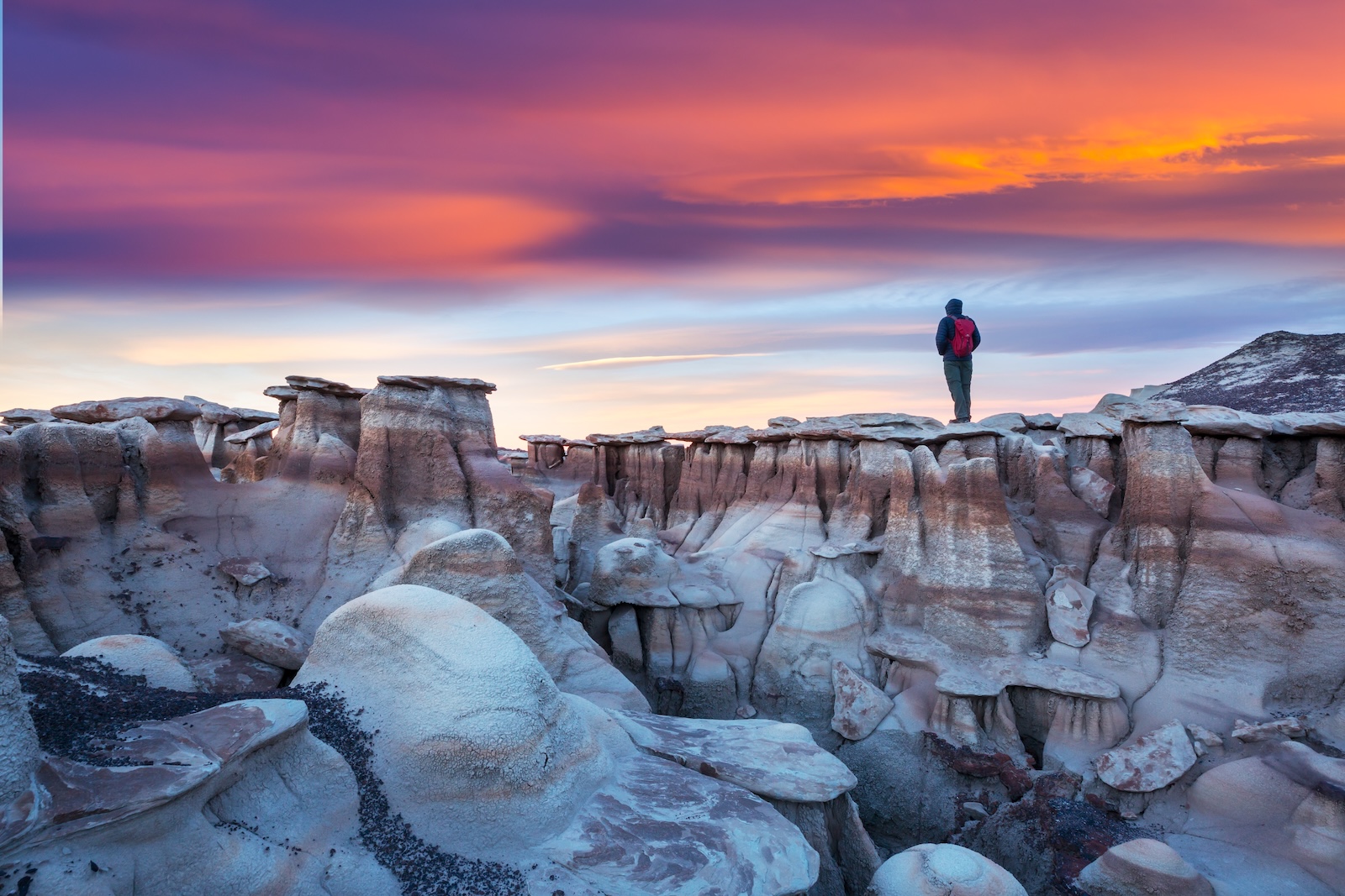 A desert landscape of rock formations with a purple and orange sky and a man hiking with a backpack at the Bisti Badlands in the De-na-zin wilderness area in New Mexico.